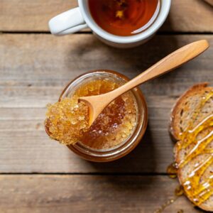 Overhead flat lay composition on weathered wooden planks showing practical honey uses. Center: open jar of crystallized raw honey with wooden spoon resting across rim