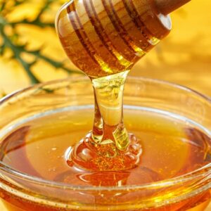 Close-up macro photograph of kenyan raw organic honey being poured from wooden honey dipper into clear glass bowl.