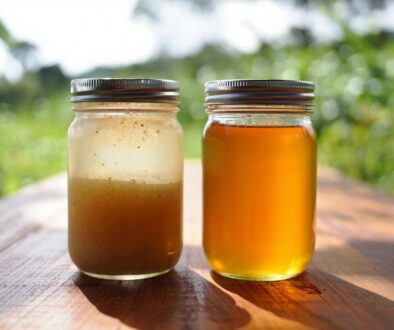 Split comparison showing two mason jars side by side on wooden surface. Left jar contains raw honey with cloudy, opaque appearance and visible fine particles settling at bottom. Right jar shows clear, transparent processed honey with uniform golden color
